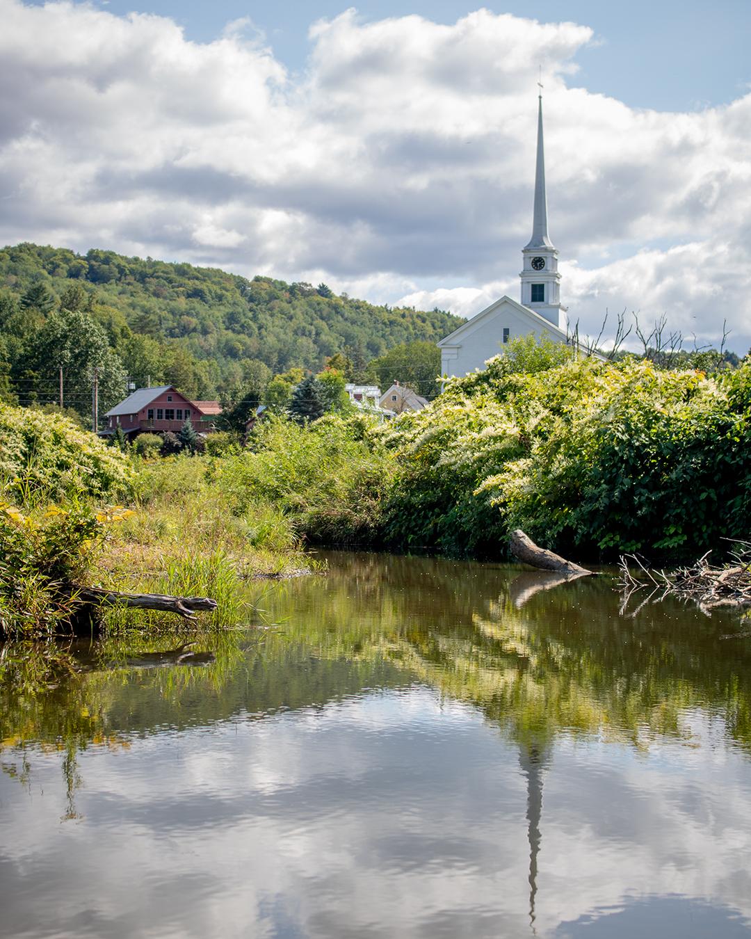 White church with a tall steeple, near a pond, surrounded by green hills and clouds.