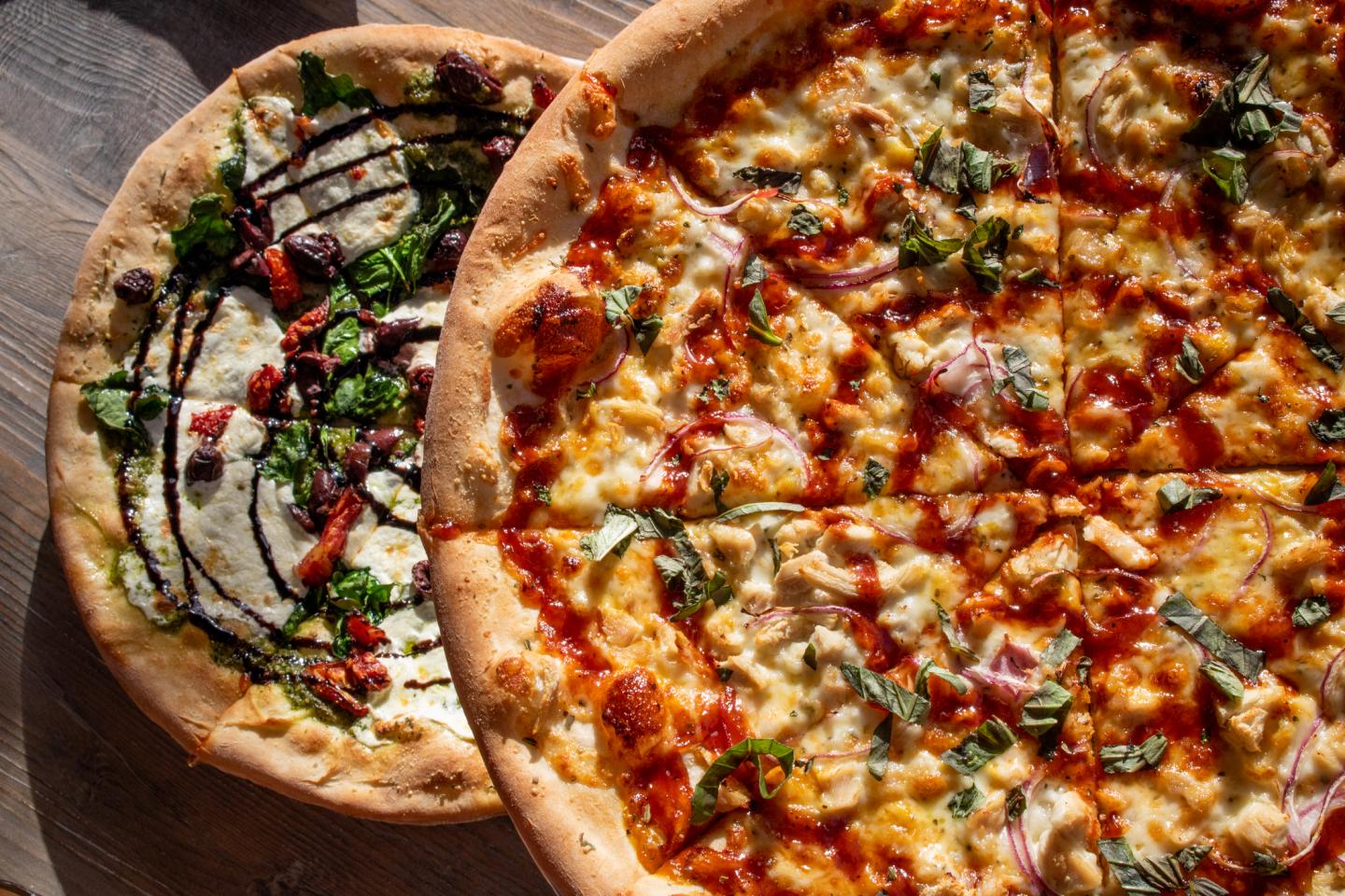Two pizzas on a wooden table, one with barbecue toppings, the other with spinach and olives.
