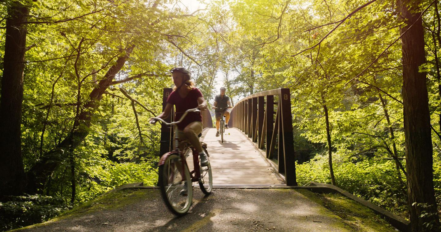 Cyclist and walker on a sunlit forest bridge.