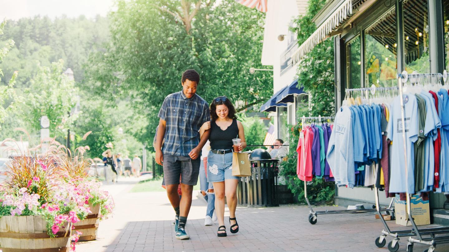 Couple walking past storefront with clothing racks on a sunny day.
