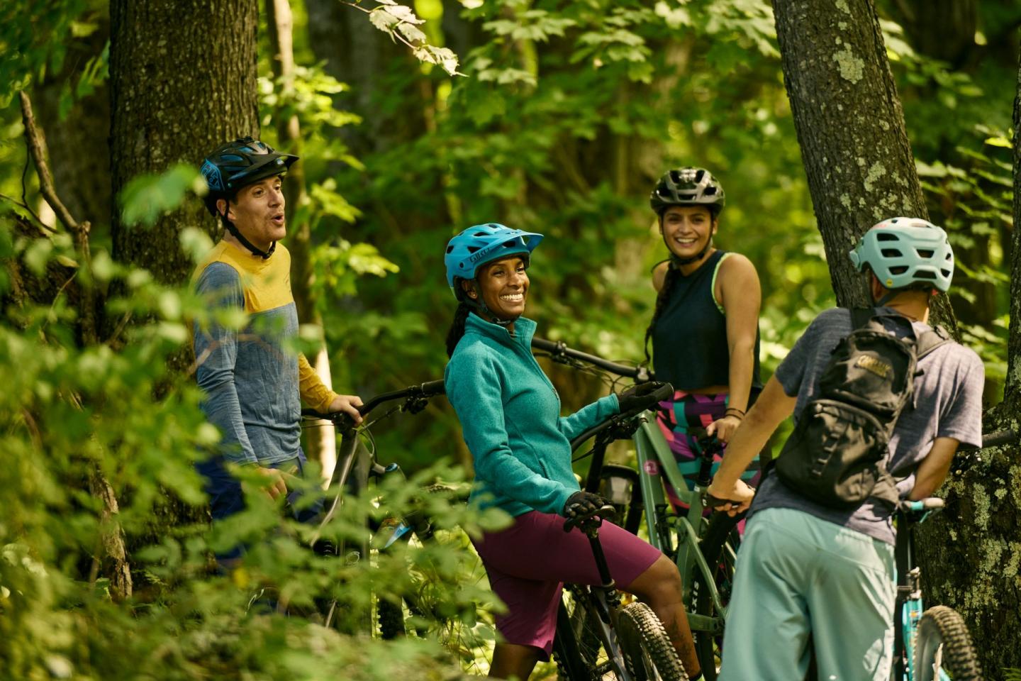 Cyclists chatting in a forest, wearing helmets and casual gear, surrounded by green trees.