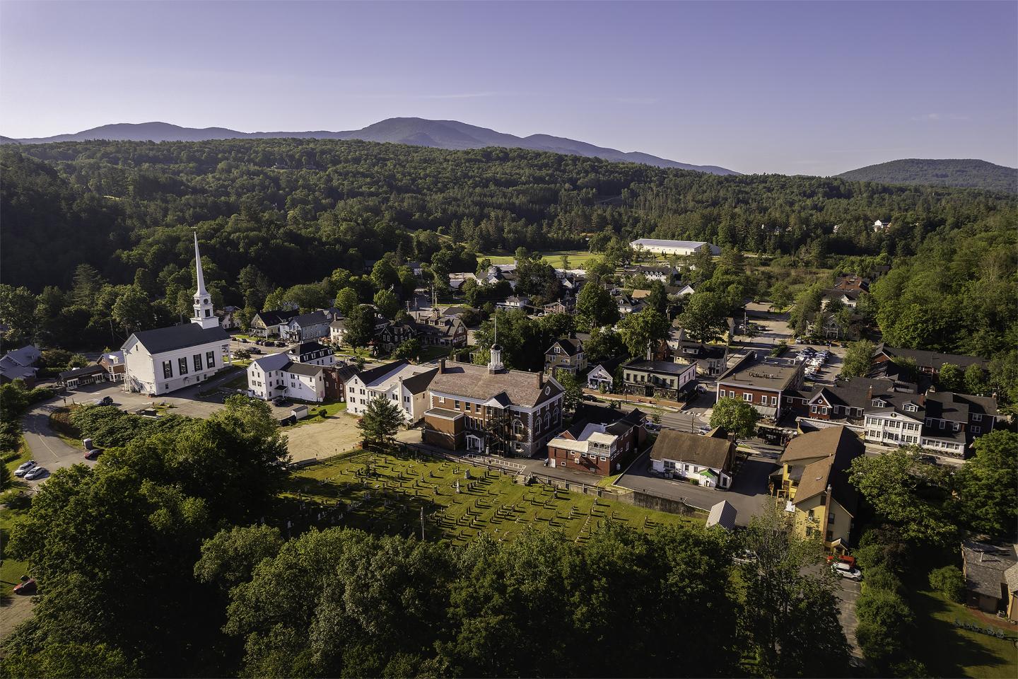 Small town with a church steeple, surrounded by trees, with mountains in the background.