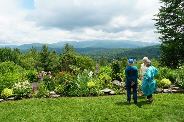 Three people in a garden overlook a mountain view under a cloudy sky.