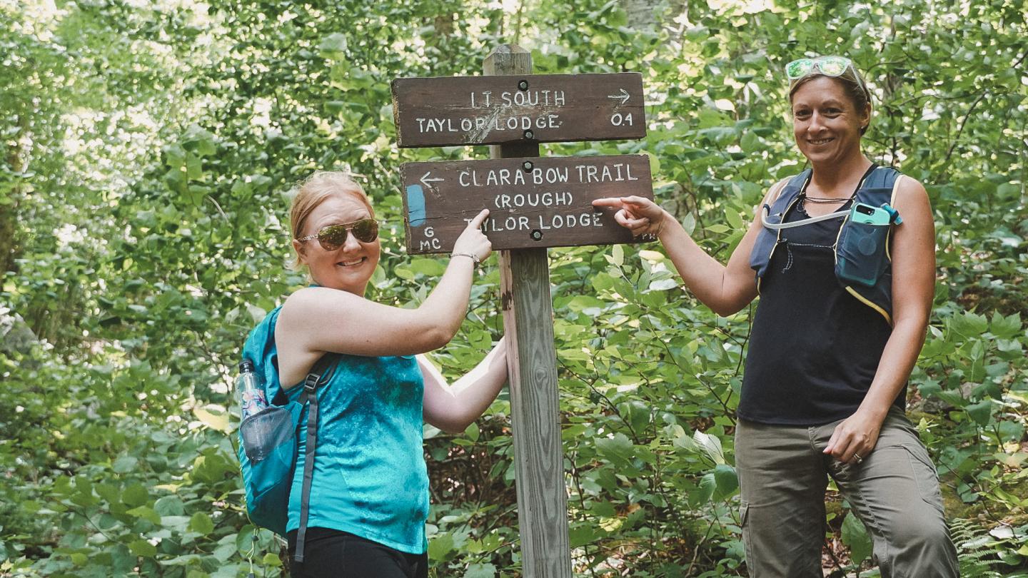 Two women point at a trail sign in a lush forest setting.