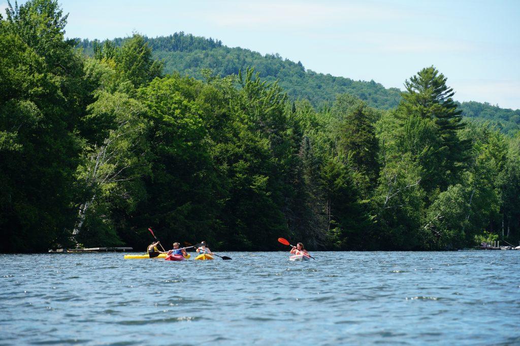 Kayakers on a calm lake surrounded by lush green trees and distant hills.