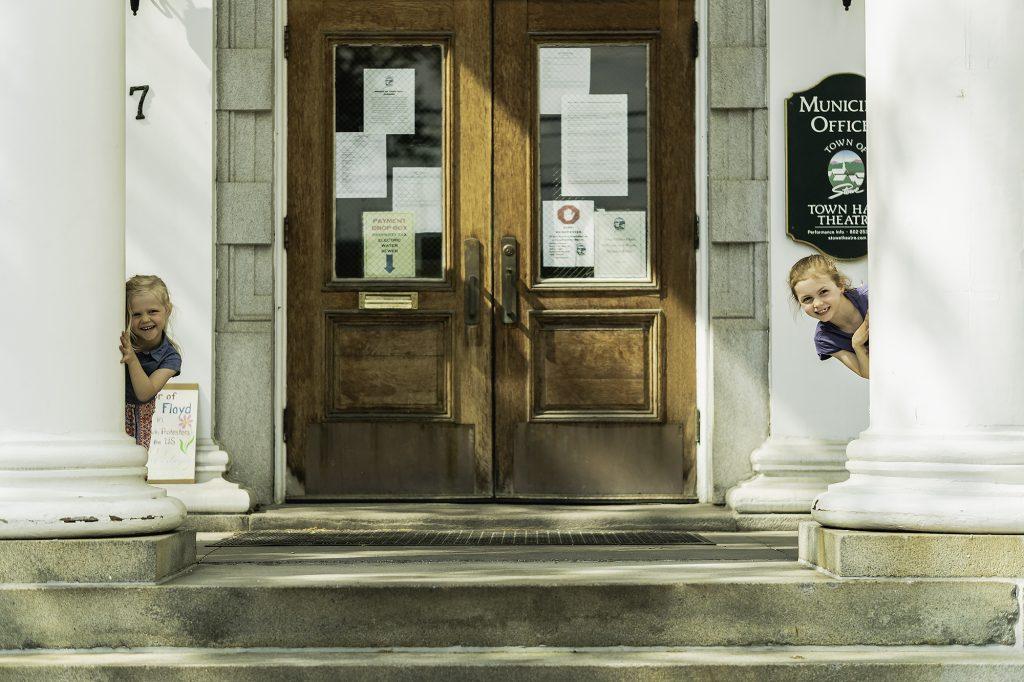 Children peeking from behind columns in front of a building entrance.