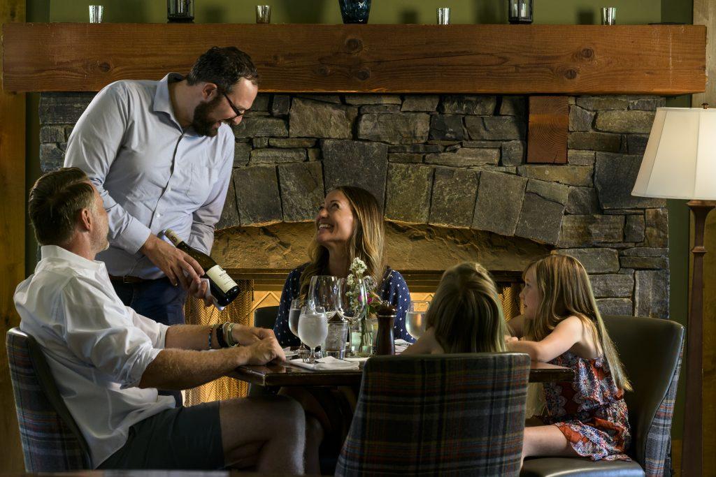 Family dining by a stone fireplace, server pouring wine.