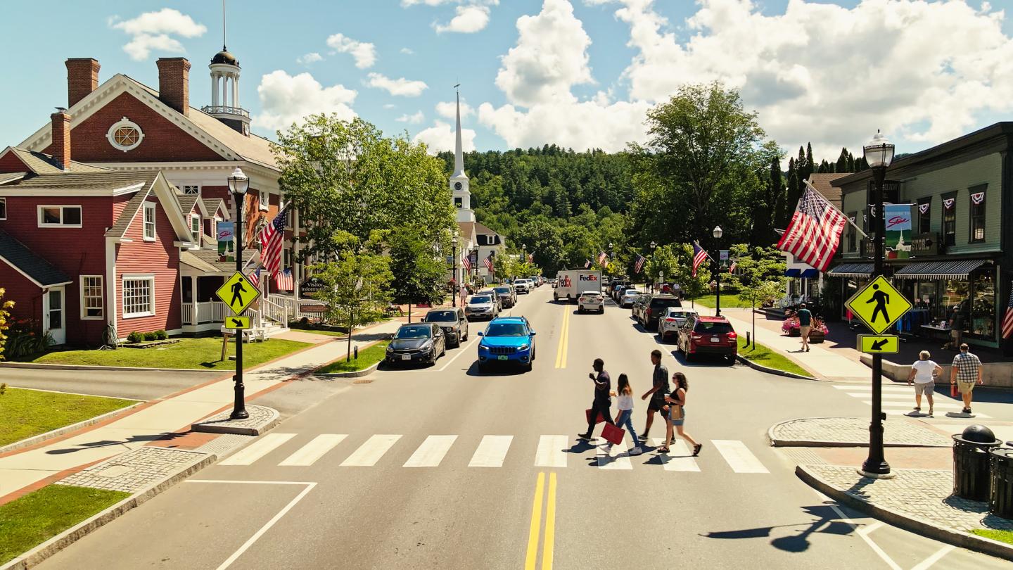 Small town street with pedestrians crossing, American flags displayed, and cars parked.