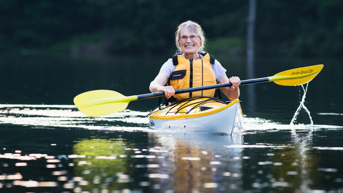 Smiling person kayaking on a calm lake, wearing a yellow life vest.