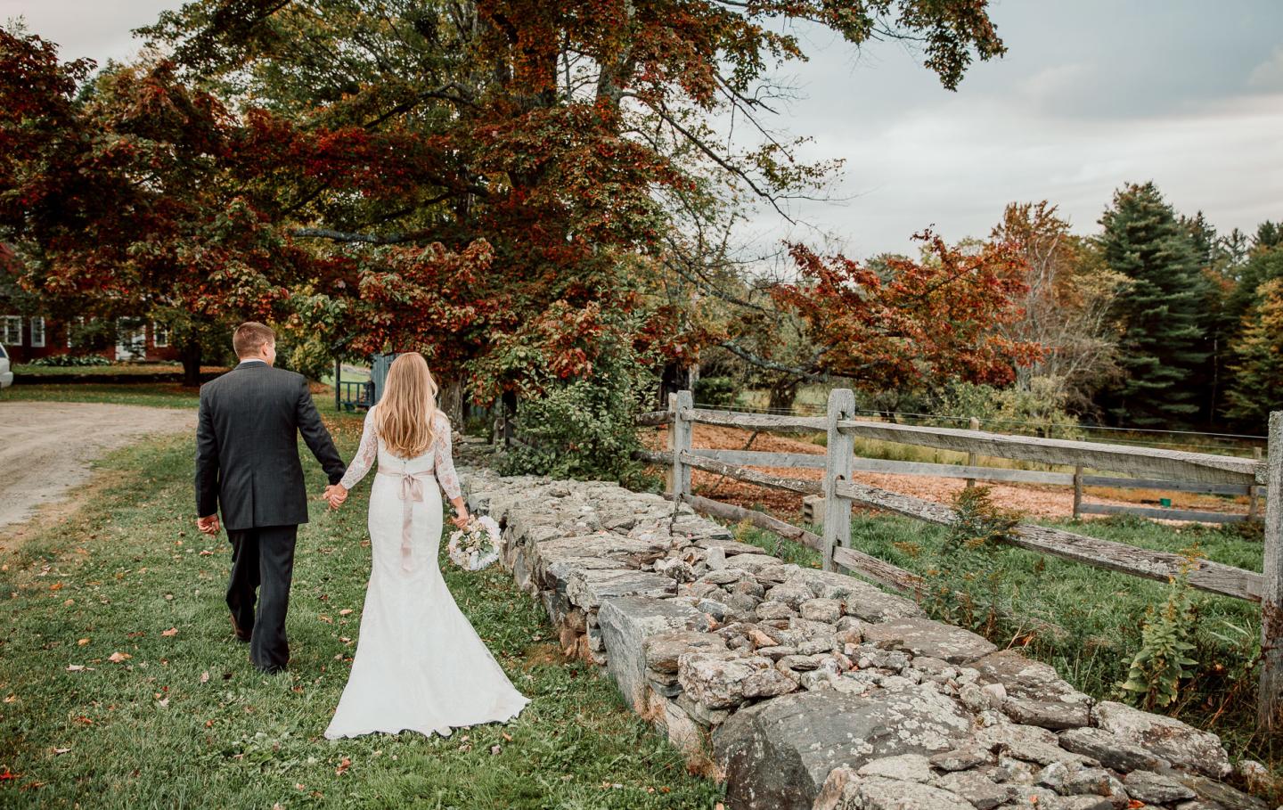 Bride and groom walk hand in hand along a rustic stone wall, surrounded by autumn foliage.