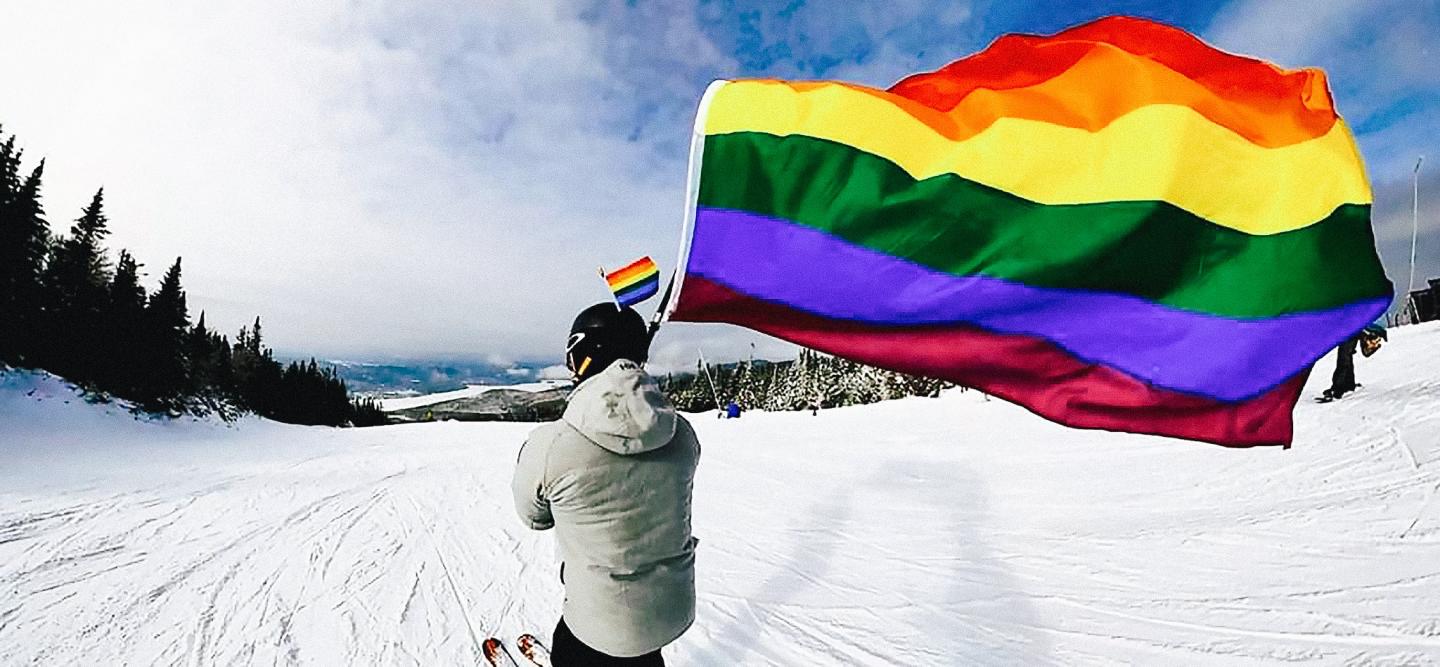 Skier holding a rainbow flag on a snowy slope.