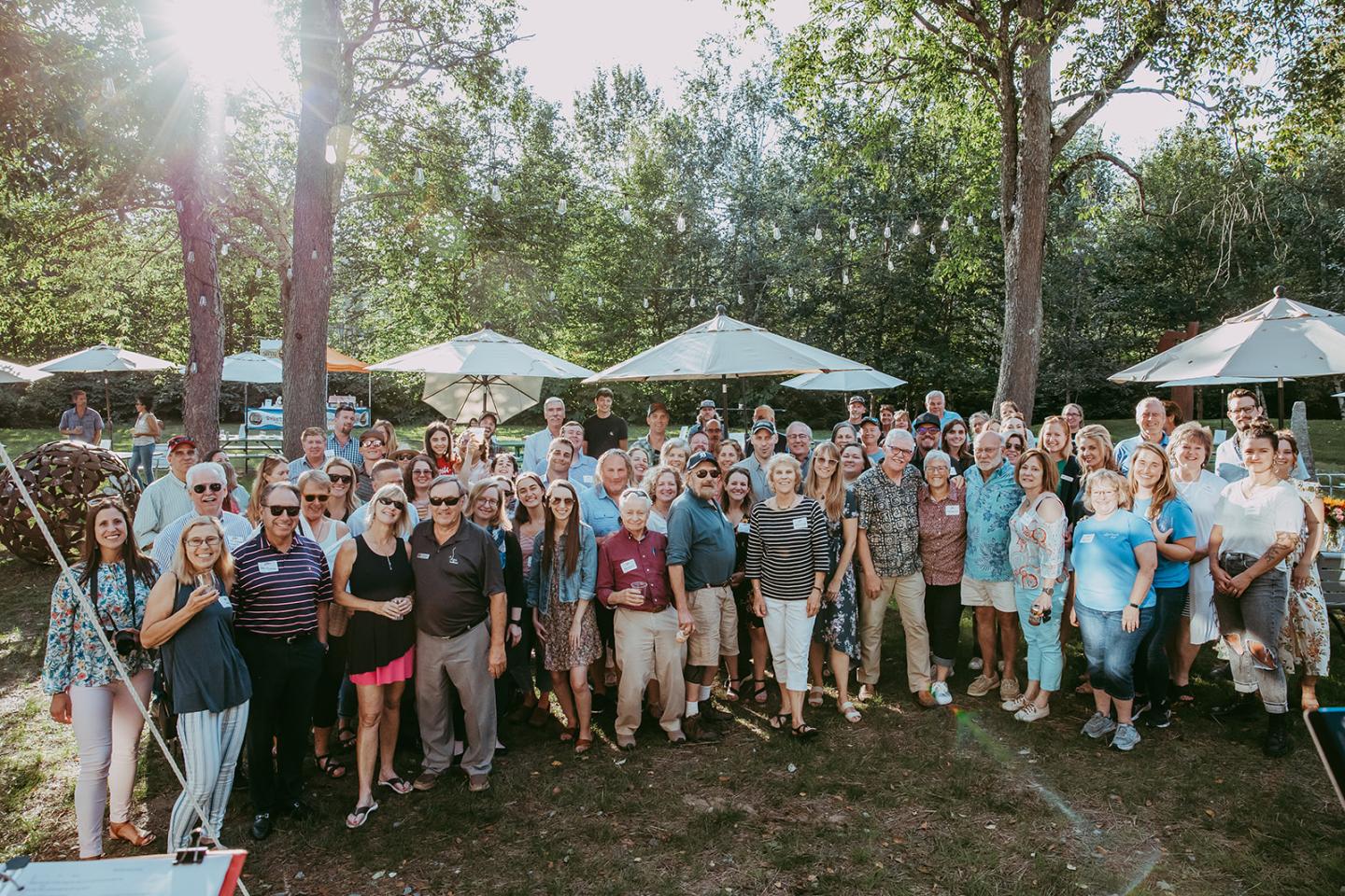 Group photo outdoors with many people under trees and parasols, sunlight filtering through.