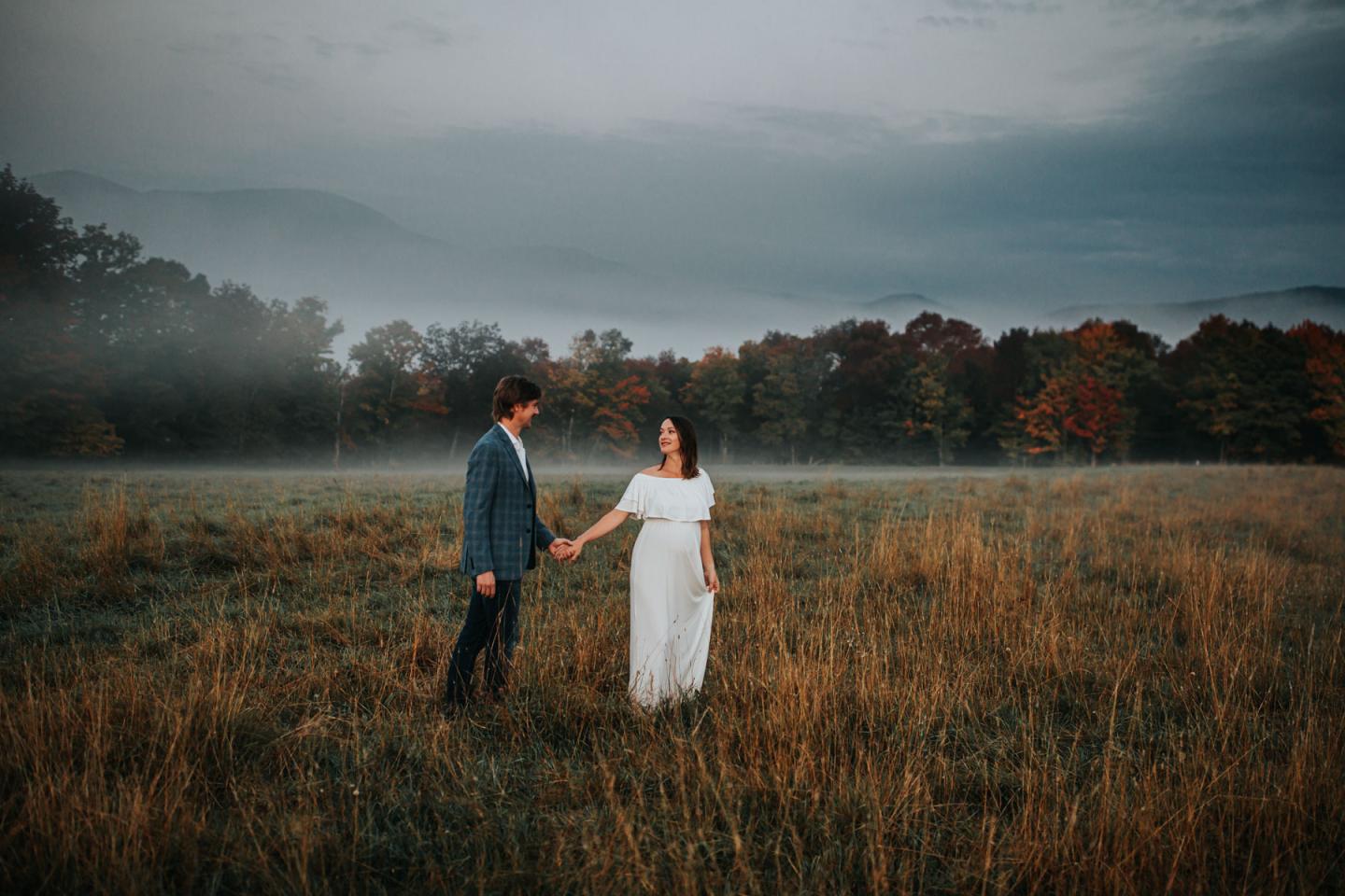 Couple holding hands in a misty field, autumn trees in the background.