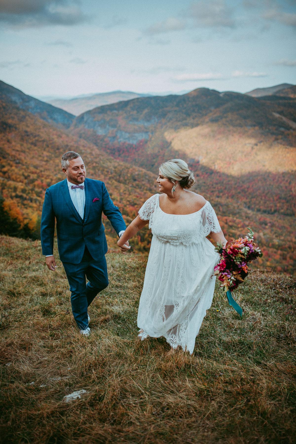 A couple walks hand in hand on a hill with mountains in the background.