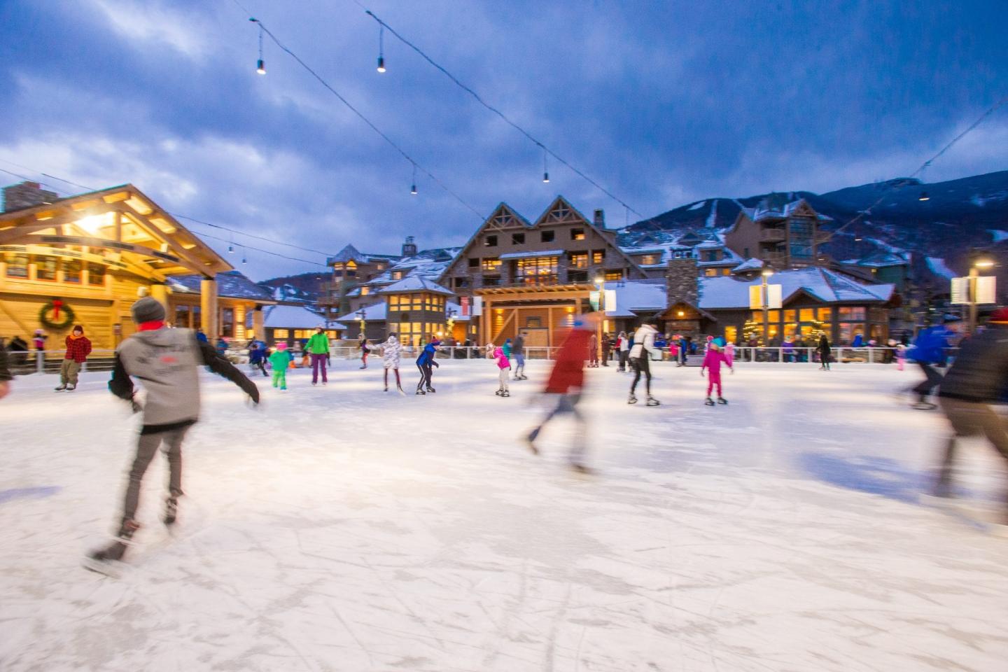 People ice skating in a village square at dusk, with lights and snow-covered buildings.