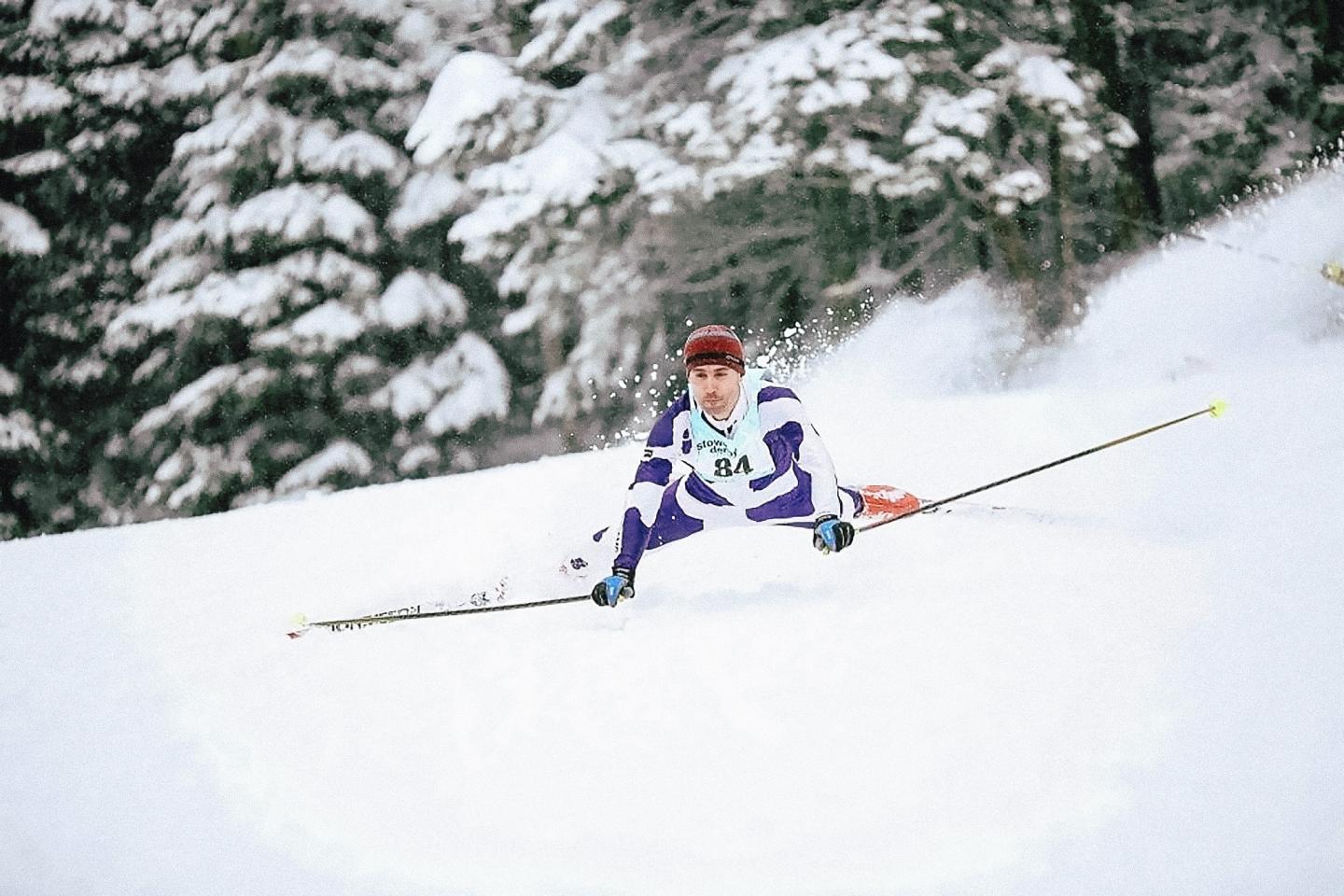 Skier in a white and purple suit racing down a snowy slope with trees in the background.
