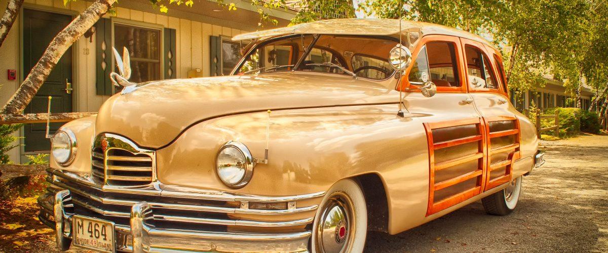 Vintage beige car in a sunlit driveway, surrounded by greenery.
