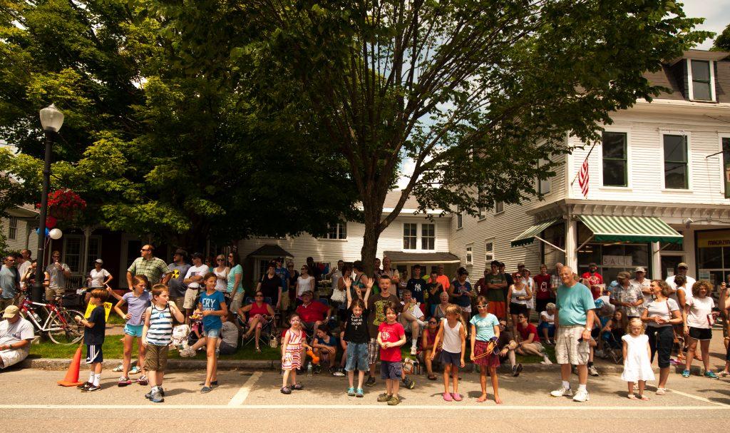 Crowd gathered on a street, under a large tree, with houses in the background.