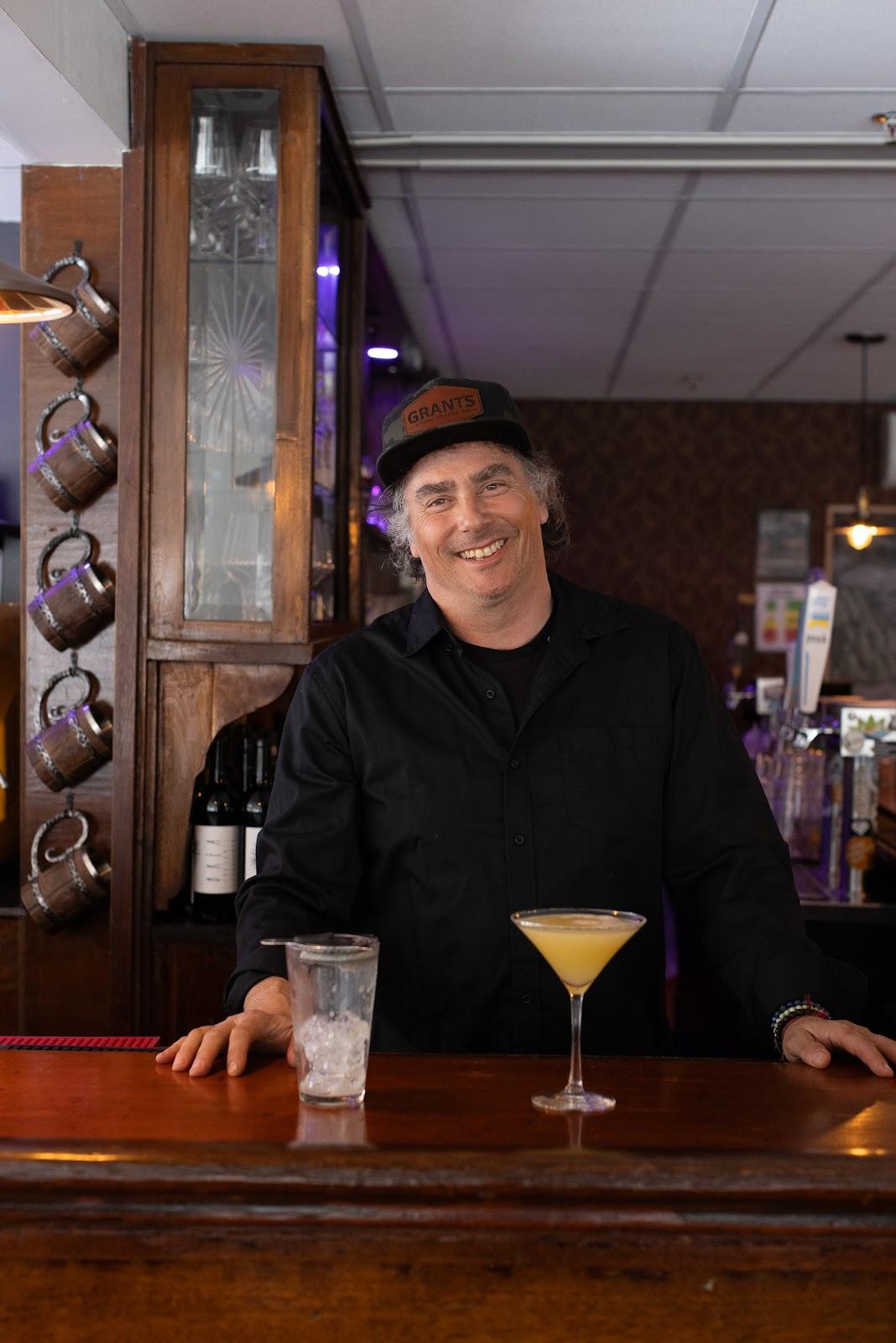 Smiling bartender with a cocktail at a wooden bar.