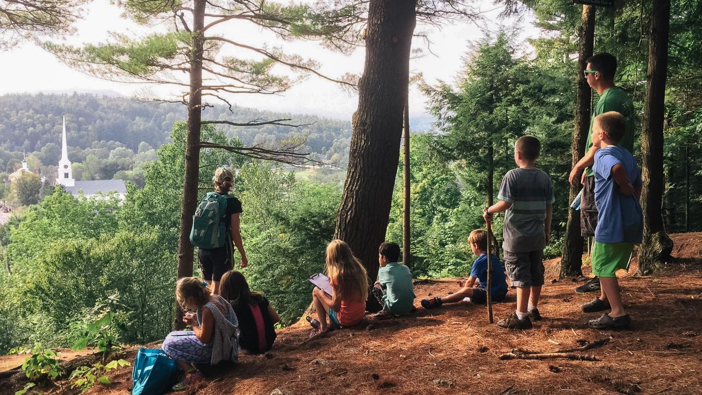 Children and an adult pause in a forest overlooking a distant church steeple.