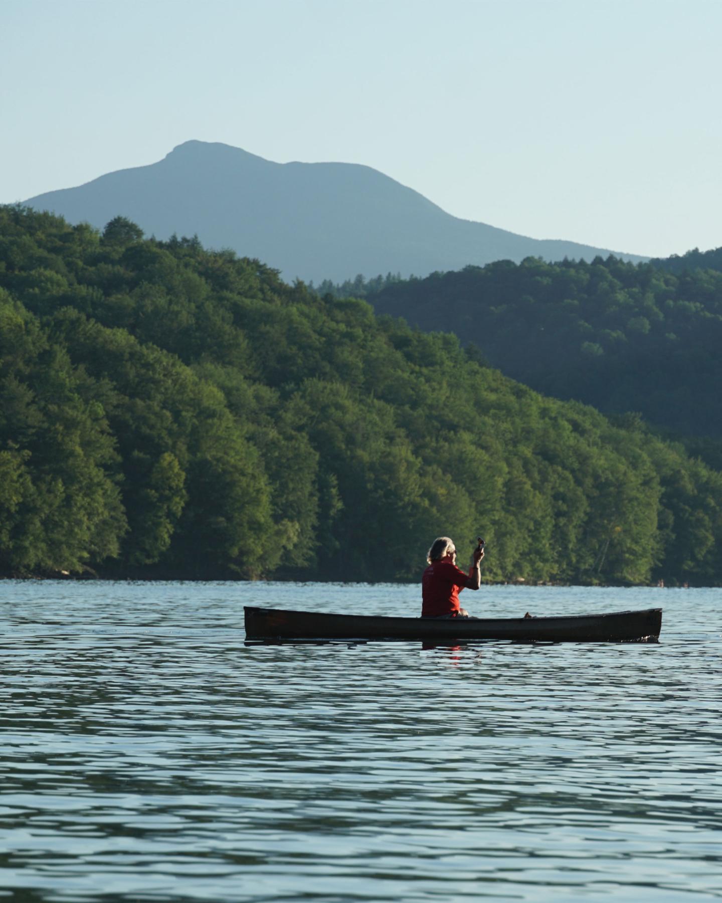 Person canoeing on a lake with forested hills and mountains in the background.