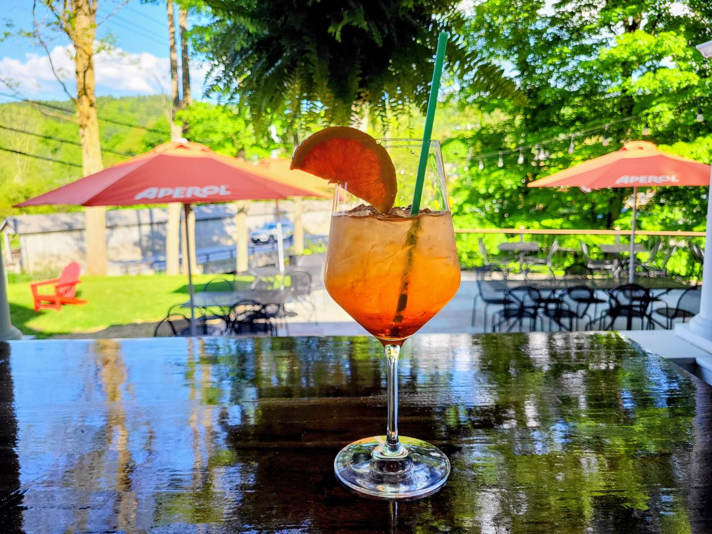 Orange cocktail with straw and citrus slice on outdoor table, sunny patio in background.