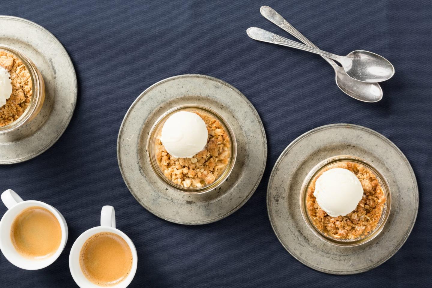 Desserts with ice cream scoops on metal plates, two coffee cups, and spoons on a dark cloth.