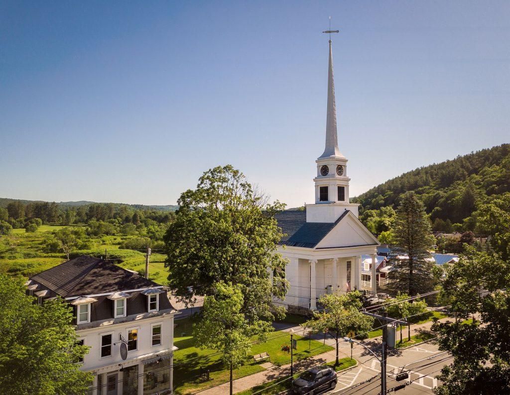 White church with tall steeple in a lush, green landscape under a clear blue sky.