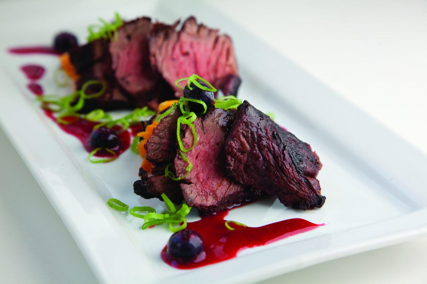 Sliced grilled steak with garnish and sauce on a white plate.