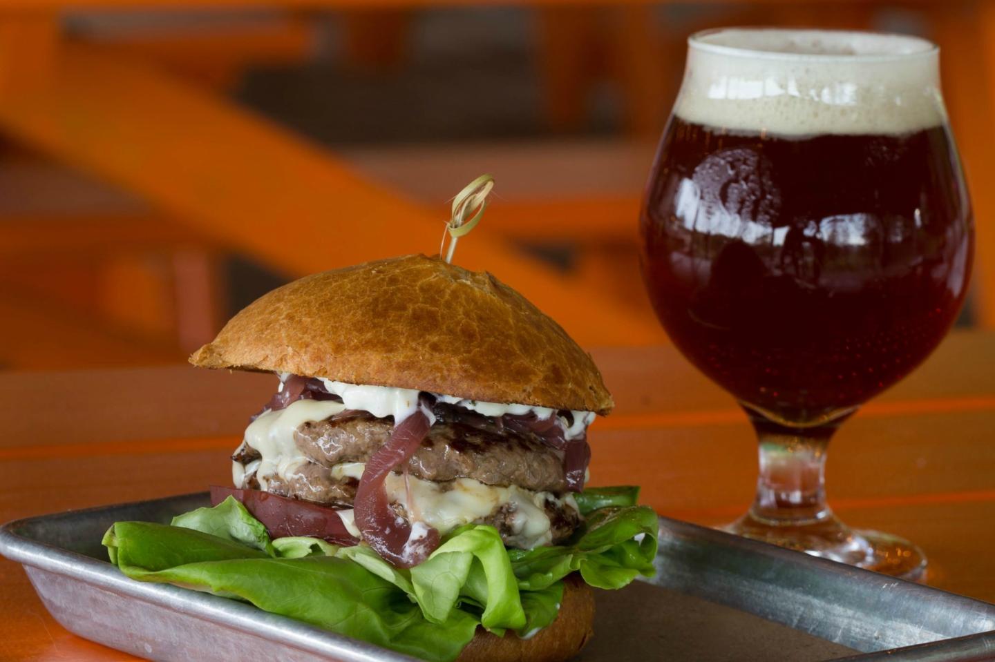 Burger with greens on a tray beside a glass of dark beer on a wooden table.