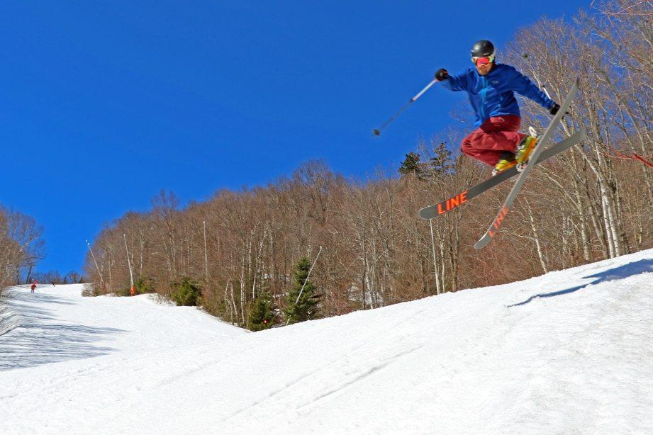 Skier in mid-air jump on a snowy slope, clear blue sky.