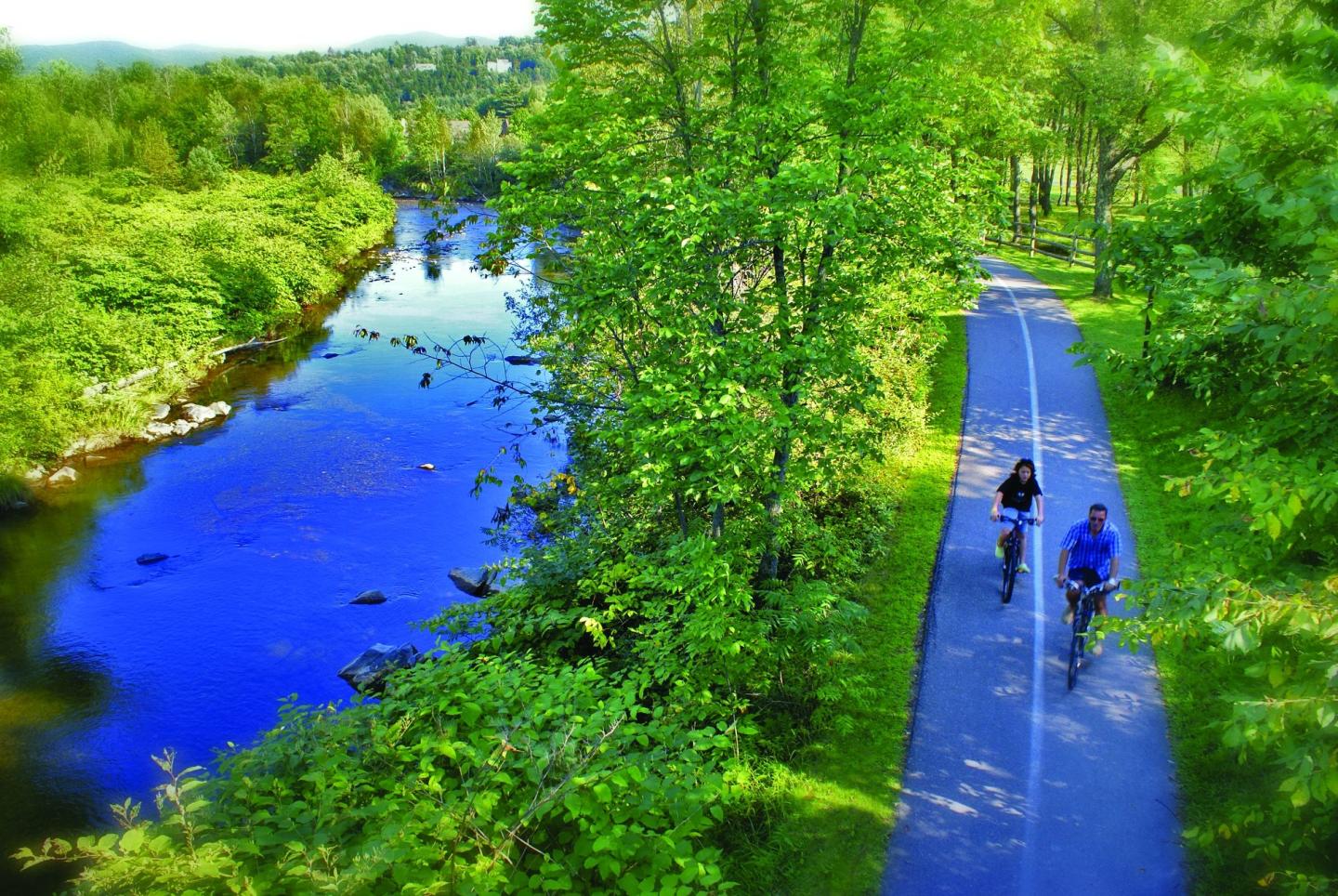 Two cyclists on a path beside a blue river, surrounded by green trees.