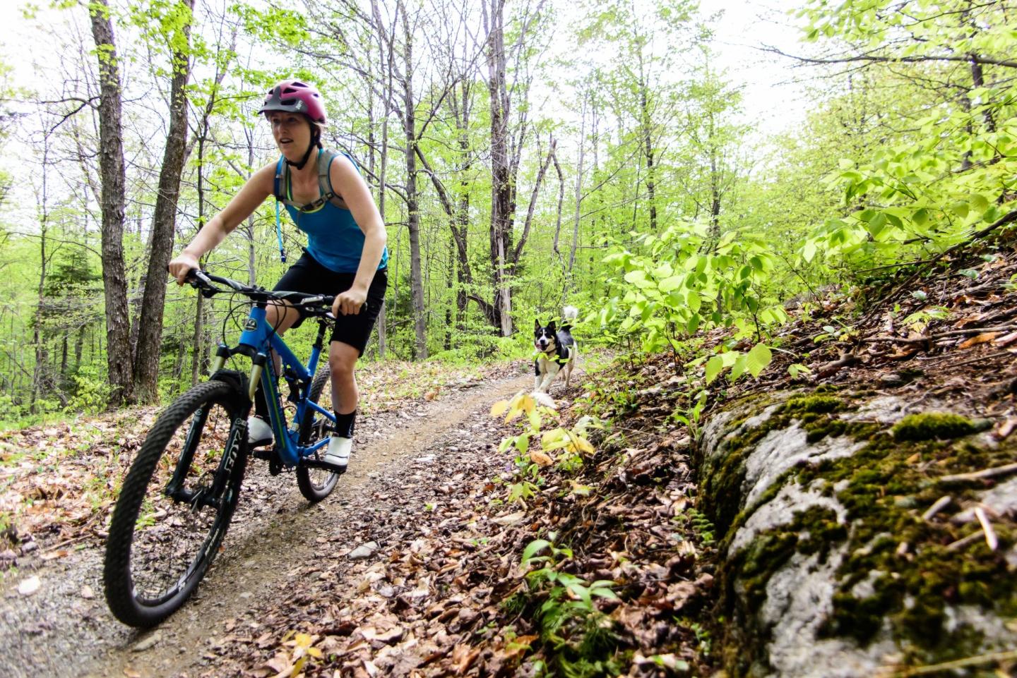 Woman biking on a forest trail, wearing a helmet and tank top.