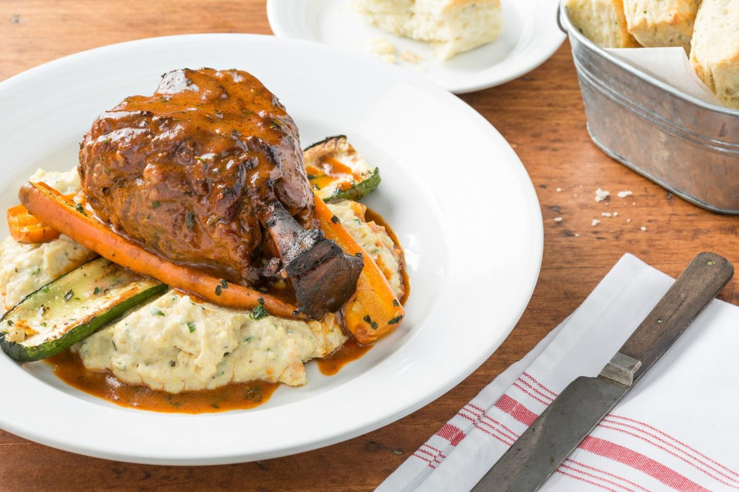 Braised lamb shank with mashed potatoes, carrots, and zucchini on a white plate.