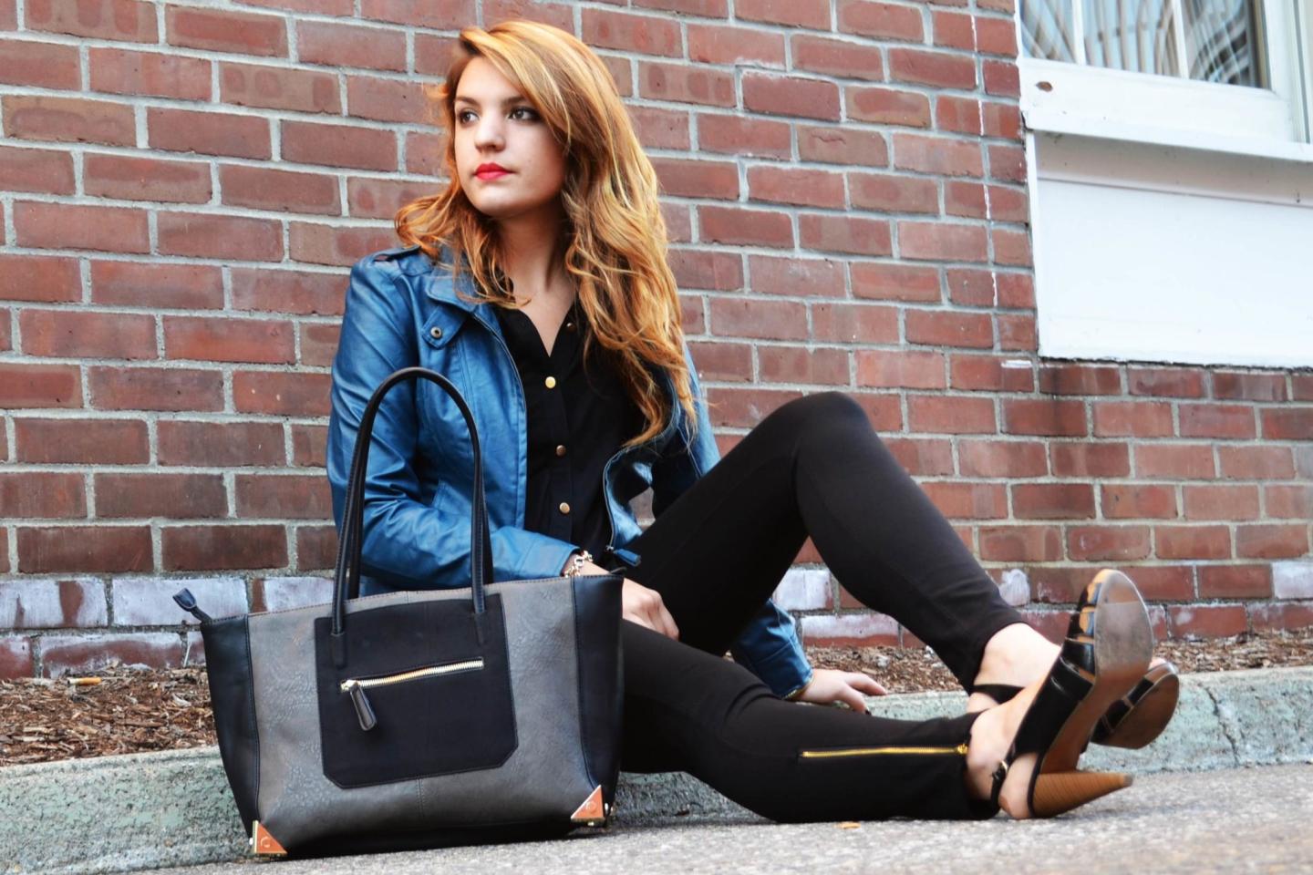 Woman sitting against brick wall with blue jacket and large gray handbag.