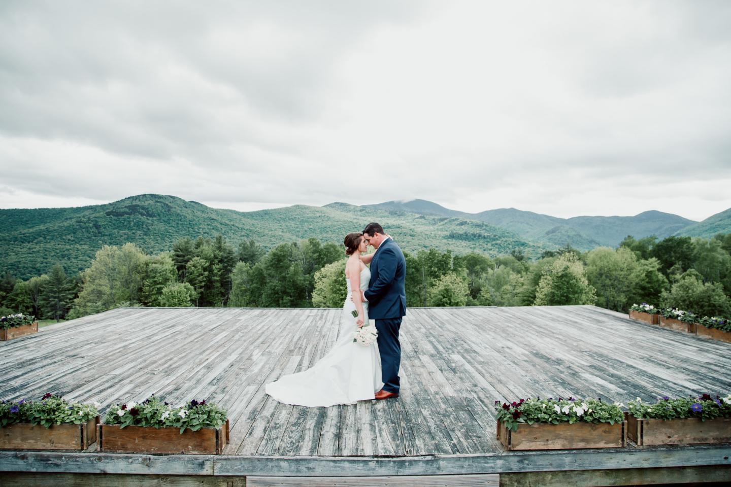 Bride and groom embrace on wooden deck with mountain backdrop.