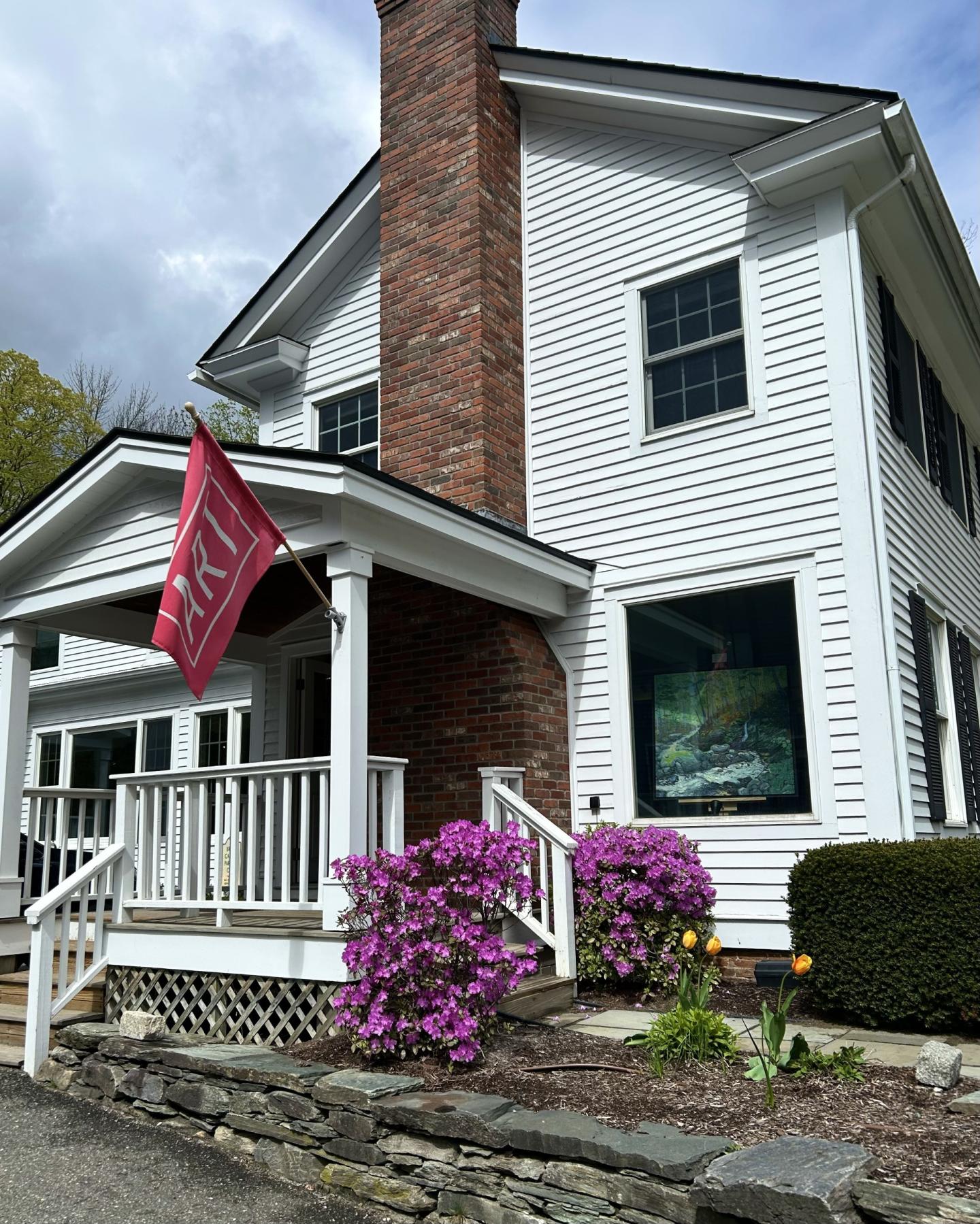 White house with a red flag, purple flowers, and a brick chimney.