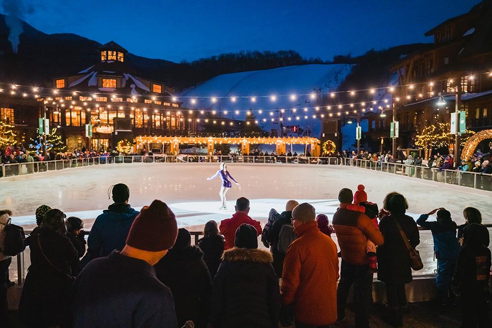 Skater performing on illuminated outdoor rink at night, surrounded by spectators.