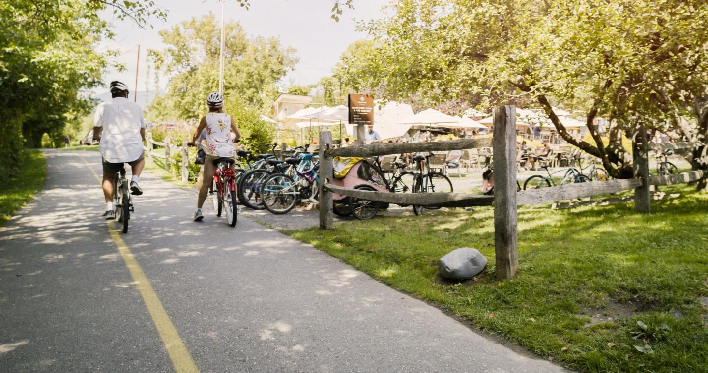 Cyclists on a sunny path near parked bikes and grassy area.