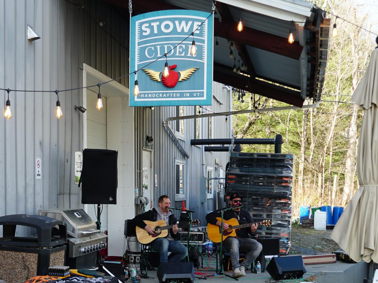 Two musicians play guitars outside under string lights near a cider sign.