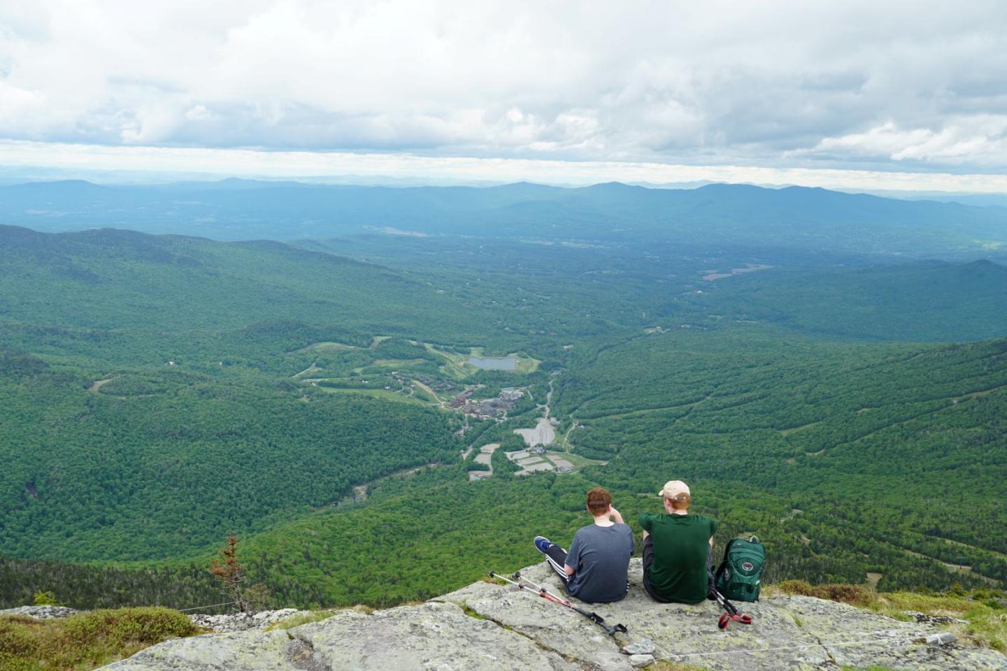 Two people sitting on a cliff, overlooking a vast green valley.
