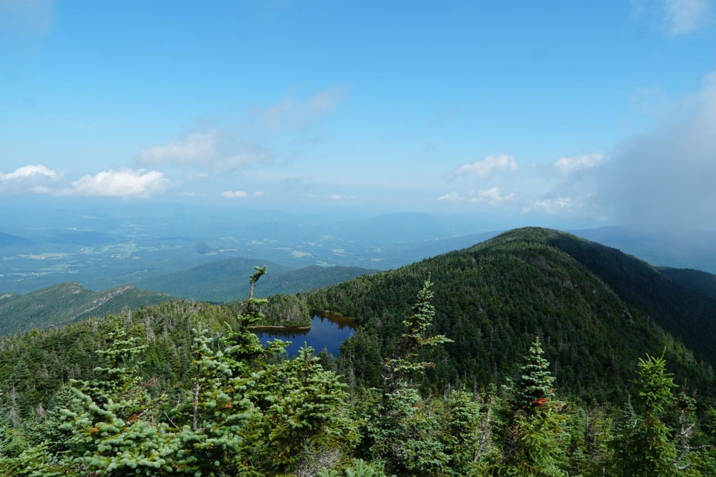 Mountain landscape with blue sky, scattered clouds, and a small lake surrounded by trees.