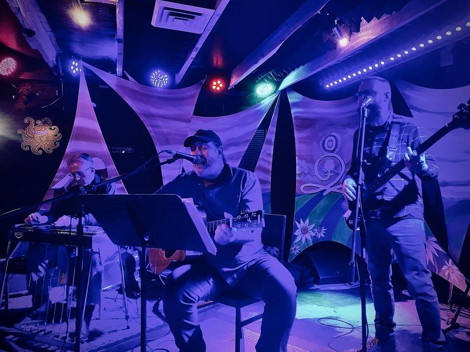 Three musicians perform on stage under colorful lights.