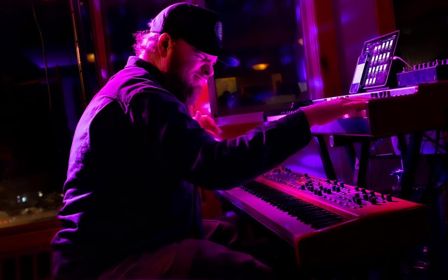 Musician playing a keyboard in dim pink and purple lighting.