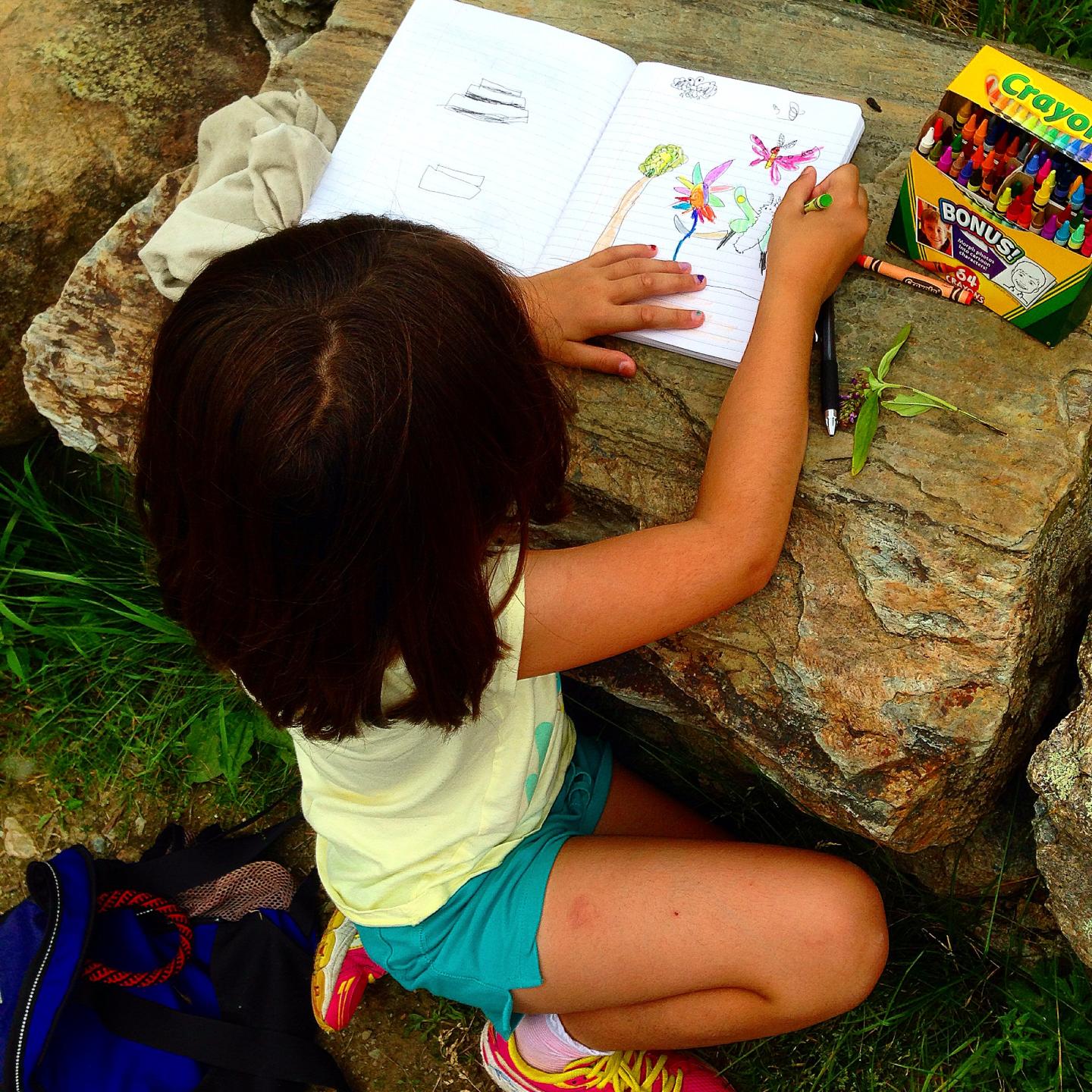 Child sitting on rocks, coloring in a book with crayons.