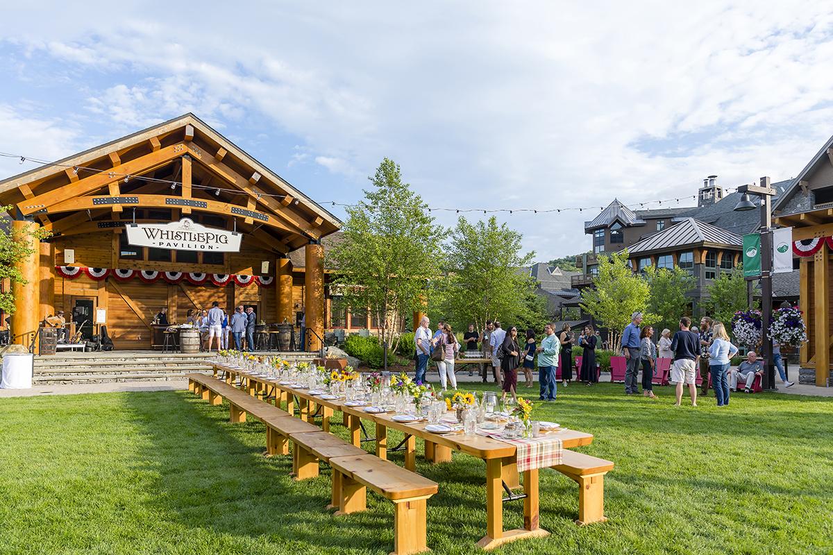 Outdoor event with wooden stage and long picnic tables on green lawn.