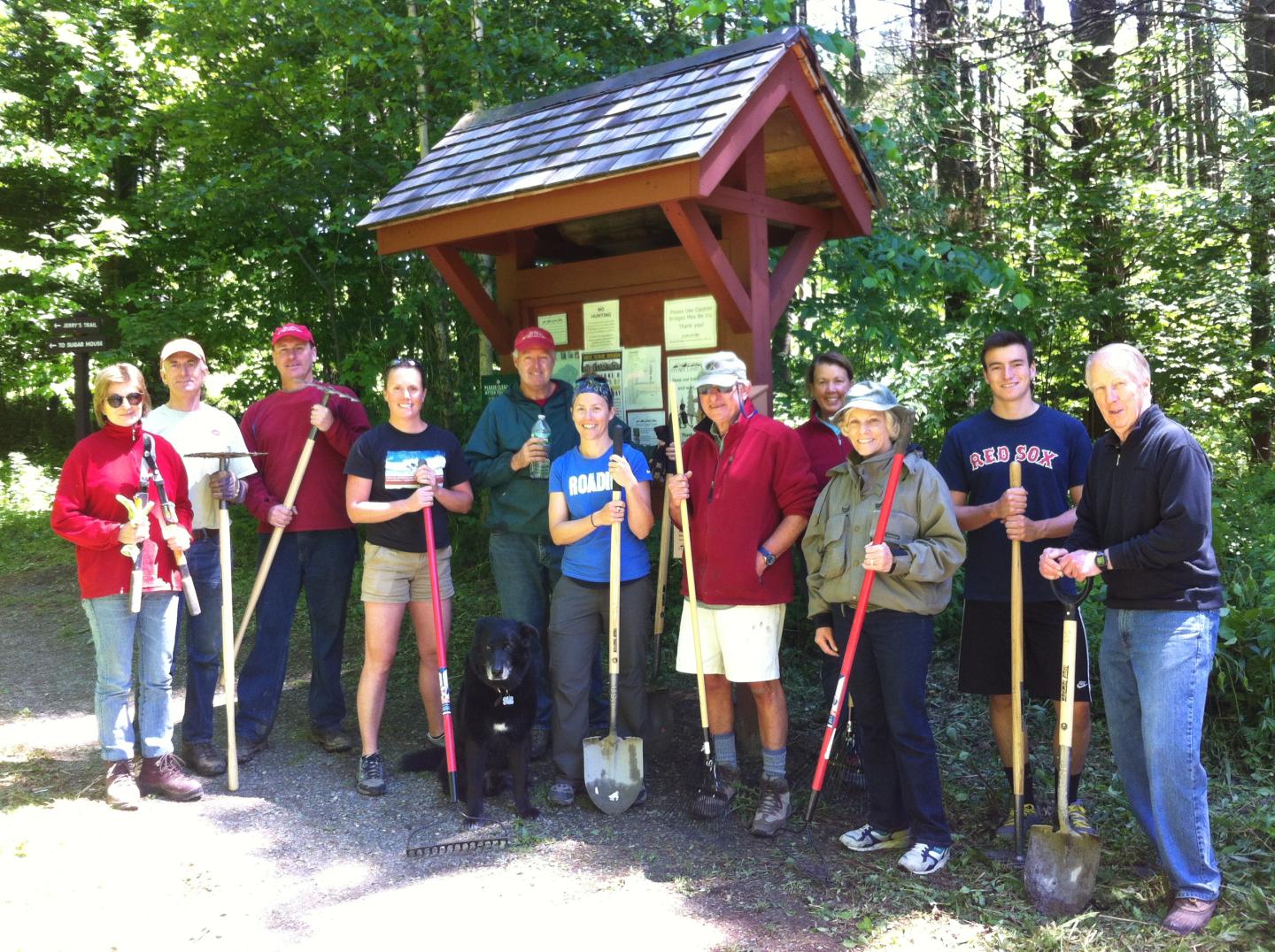 Group with shovels and dog by wooded trail sign.