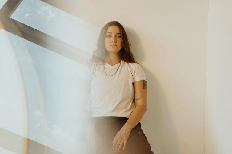 Woman in a white shirt leans against a wall, soft light effect.