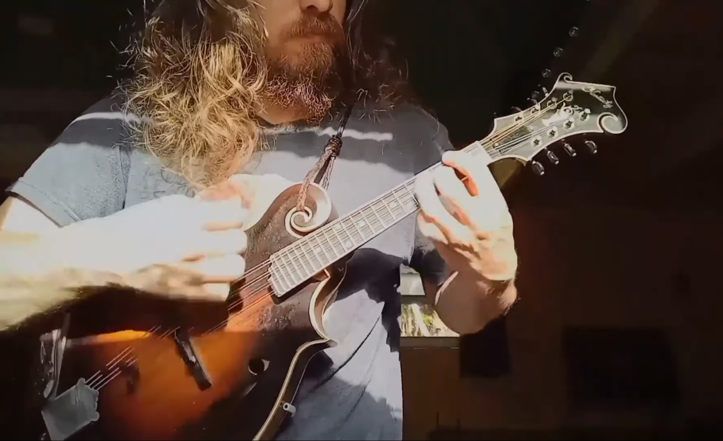 Long-haired person playing a mandolin in a sunlit room.