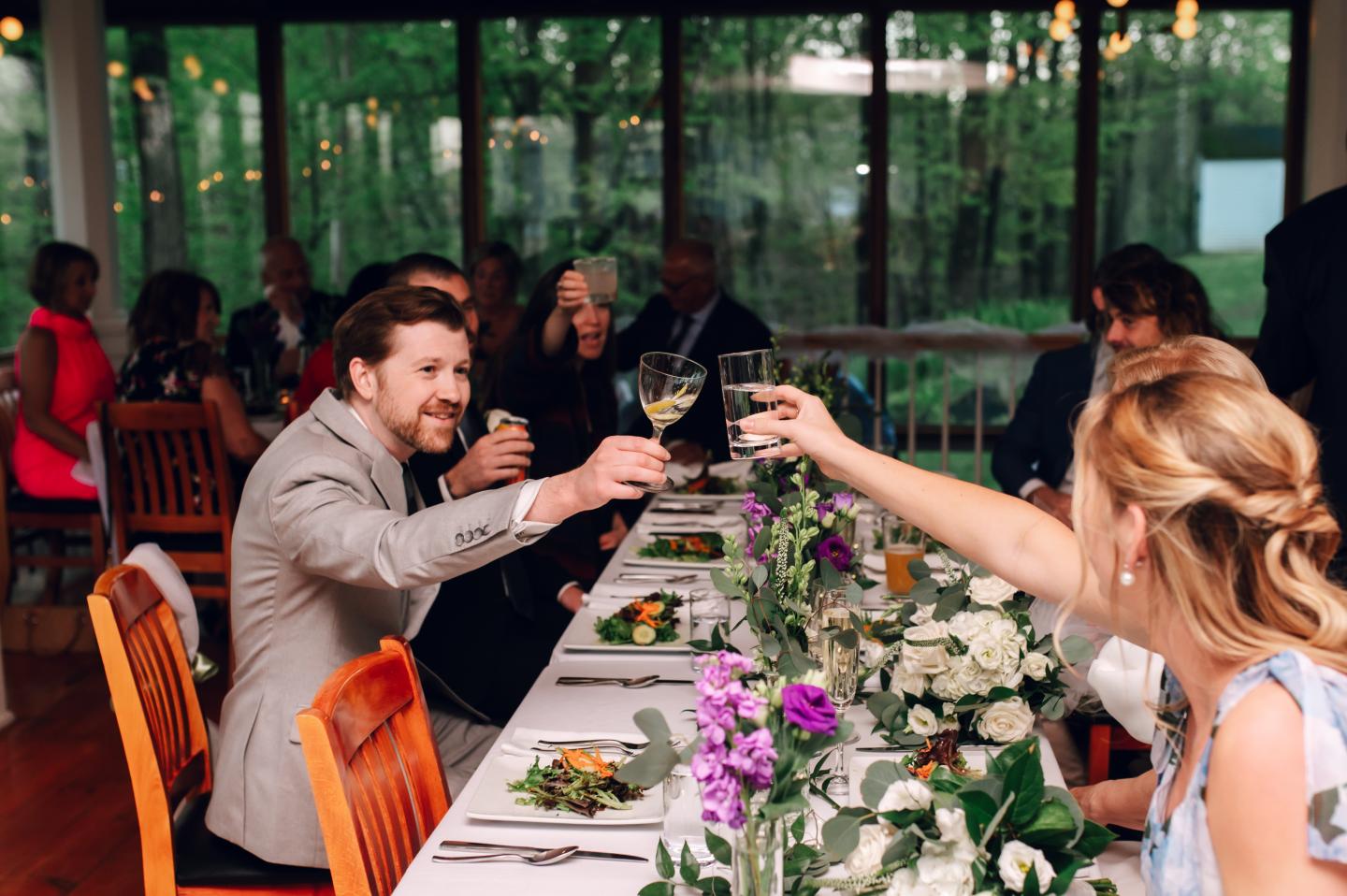 Dining table toast with smiling guests, floral centerpiece, and lush green backdrop.